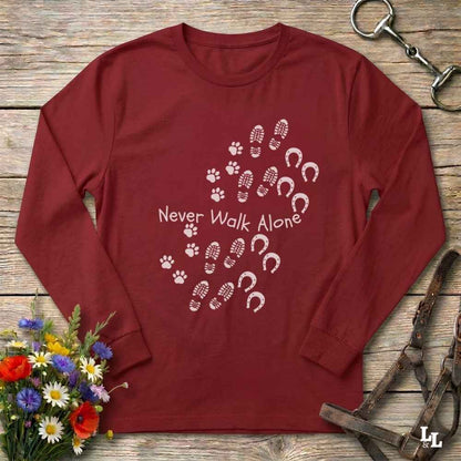 A long-sleeve crewneck t-shirt in maroon red, laid flat on a rustic wooden surface. The shirt features a white graphic print of horse hooves and paw prints arranged in a heart shape around the text 'Never Walk Alone'.