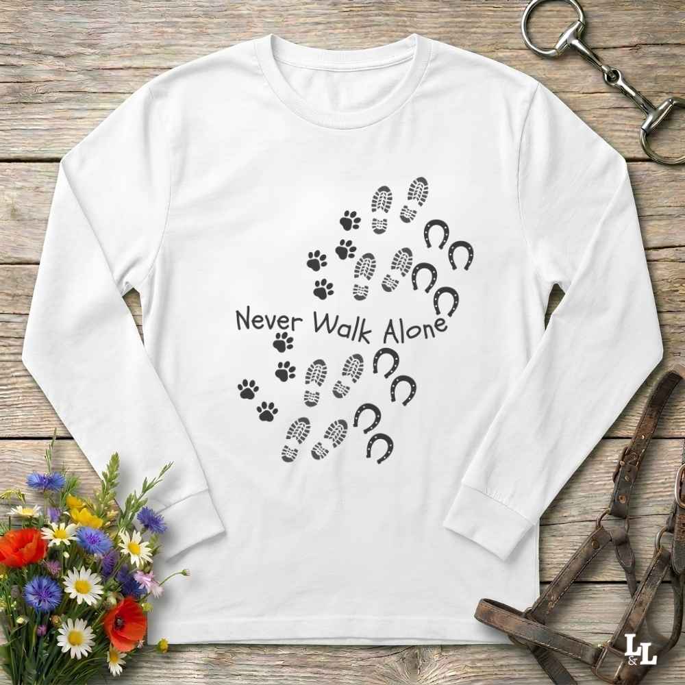 A white long-sleeve crewneck shirt laid flat on a rustic wooden surface, featuring a black graphic of paw prints, hoof prints, and the text 'Never Walk Alone.' Decorative flowers and horse tack are placed around the shirt as styling props.
