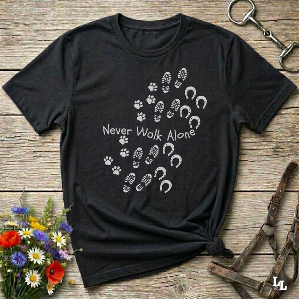 A black short-sleeve graphic T-shirt laid flat on a rustic wooden surface, with white paw prints, hoof prints, and the text 'Never Walk Alone' across the chest. Decorative flowers and horse tack are placed around the shirt.