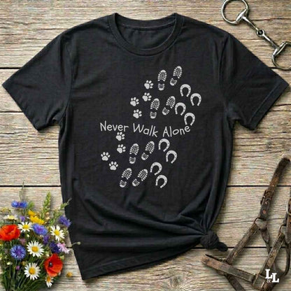 A black short-sleeve graphic T-shirt laid flat on a rustic wooden surface, with white paw prints, hoof prints, and the text 'Never Walk Alone' across the chest. Decorative flowers and horse tack are placed around the shirt.