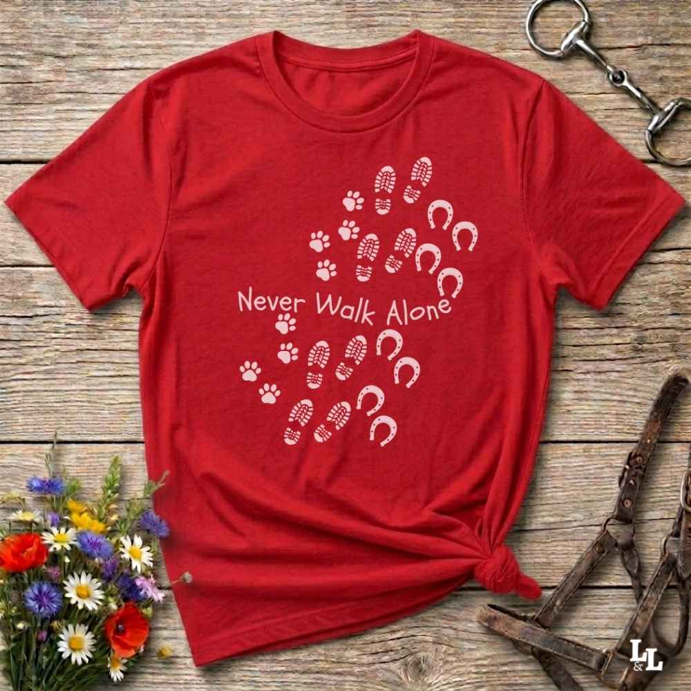A red short-sleeve T-shirt laid flat on a rustic wooden surface, printed with white paw prints, hoof prints, and the phrase 'Never Walk Alone.' Decorative flowers and horse tack are placed around it as styling props.