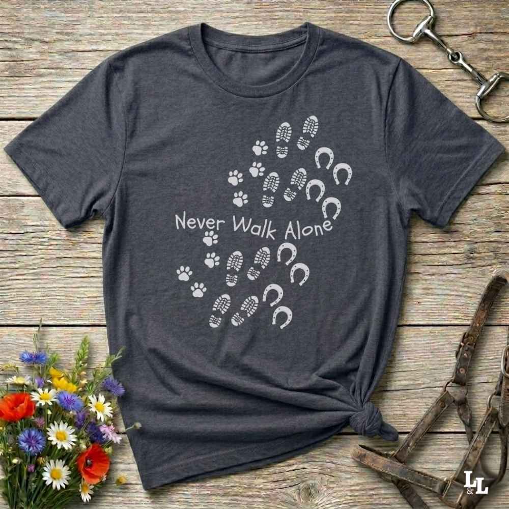 A dark heather gray t-shirt with a white graphic print of paw prints, hoof prints, and the text 'Never Walk Alone' arranged in a heart shape. The shirt is styled flat on a rustic wooden background with decorative flowers and horse tack.