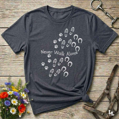 A dark heather gray t-shirt with a white graphic print of paw prints, hoof prints, and the text 'Never Walk Alone' arranged in a heart shape. The shirt is styled flat on a rustic wooden background with decorative flowers and horse tack.