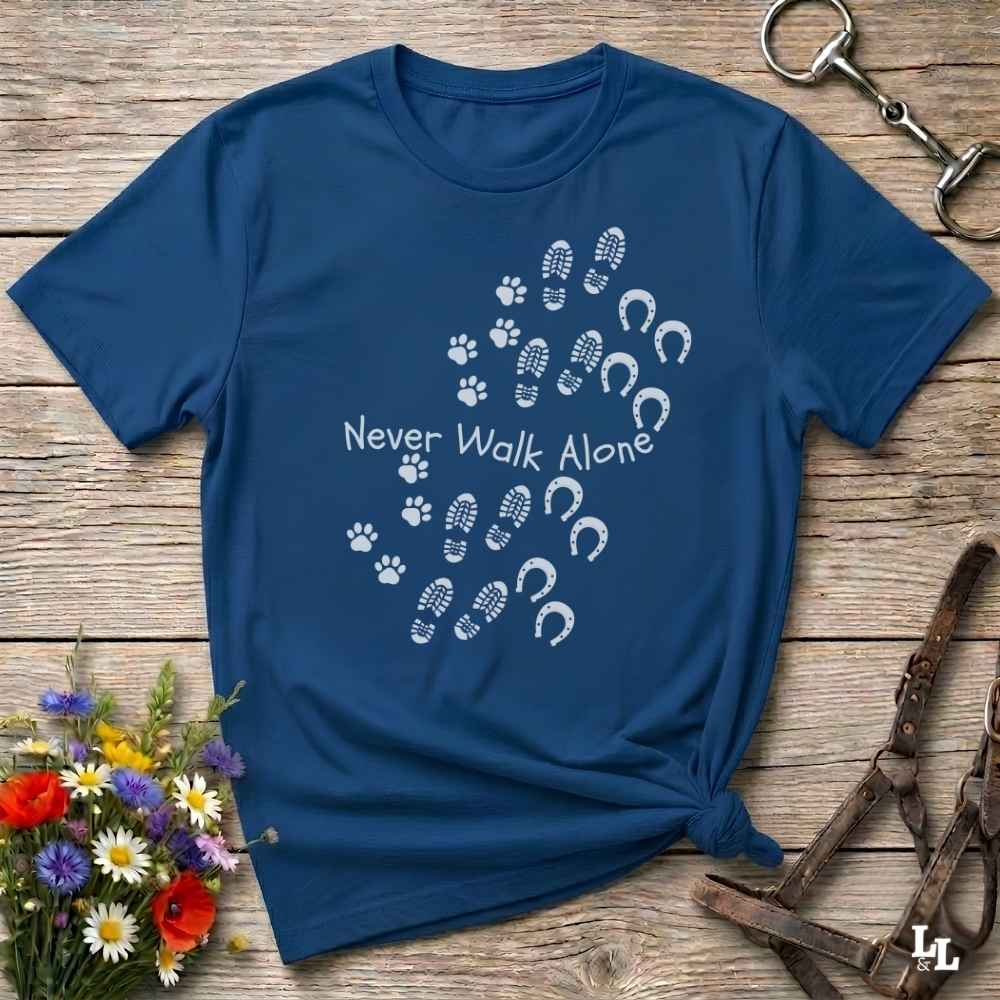 A navy blue short-sleeve t-shirt with a white graphic print reading 'Never Walk Alone' alongside paw prints, hoof prints, and footprints, styled flat on a wooden surface with equestrian accessories and flowers.