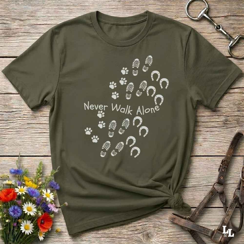 An olive green short-sleeve t-shirt with a white graphic print reading 'Never Walk Alone' accompanied by paw prints, hoof prints, and footprints, styled in a flat-lay on a rustic wooden background with equestrian accessories.