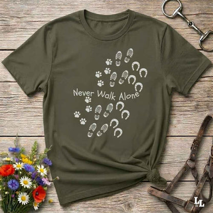 An olive green short-sleeve t-shirt with a white graphic print reading 'Never Walk Alone' accompanied by paw prints, hoof prints, and footprints, styled in a flat-lay on a rustic wooden background with equestrian accessories.