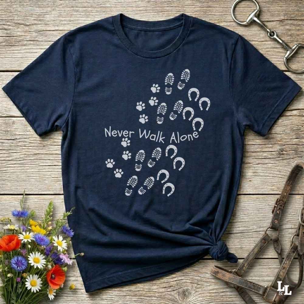 A navy blue short-sleeve T-shirt laid flat on a rustic wooden surface, printed with white paw prints, hoof prints, and the phrase 'Never Walk Alone.' The shirt is styled with flowers and equestrian tack around it.