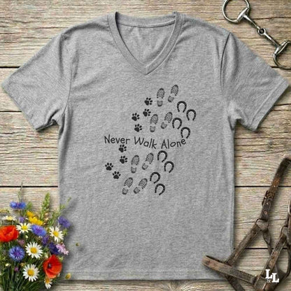 A heather gray V-neck t-shirt with a black graphic print of paw prints, hoof prints, and the text 'Never Walk Alone' arranged in a heart shape. The shirt is displayed flat on a rustic wooden background with equestrian accessories and flowers.
