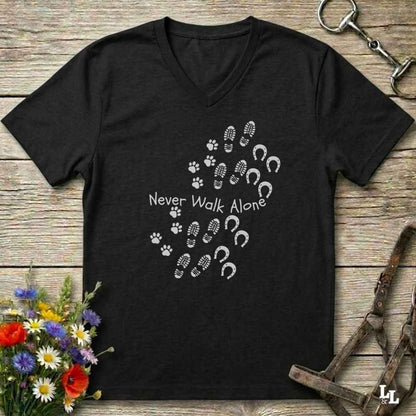 A black V-neck t-shirt with a white graphic print reading 'Never Walk Alone' accompanied by paw prints, hoof prints, and horseshoes, displayed on a rustic wooden background with equestrian accessories and flowers.
