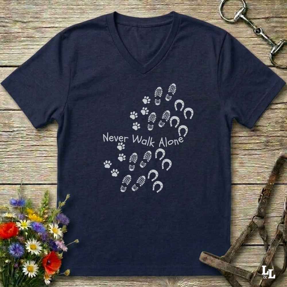 A navy blue V-neck t-shirt with a white graphic print of horse hooves and paw prints spelling 'Never Walk Alone' on a rustic wooden background, styled with equestrian tack and flowers.