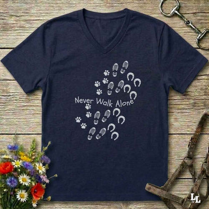 A navy blue V-neck t-shirt with a white graphic print of horse hooves and paw prints spelling 'Never Walk Alone' on a rustic wooden background, styled with equestrian tack and flowers.