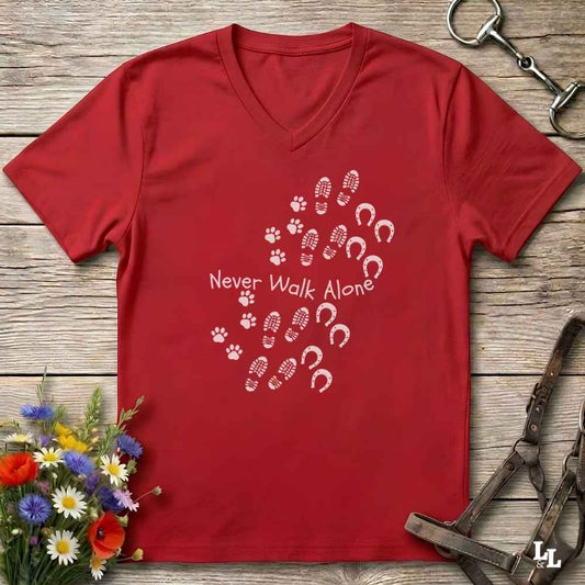 A red V-neck t-shirt with a white graphic print reading 'Never Walk Alone' accompanied by paw prints, hoof prints, and horseshoes, laid flat on a rustic wooden background with equestrian accessories and flowers.