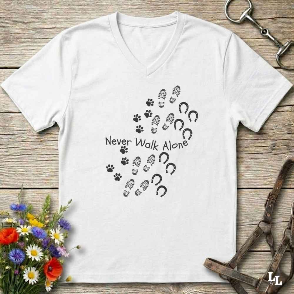 A white V-neck t-shirt with a black graphic print of paw prints, hoof prints, and the text 'Never Walk Alone' laid flat on a rustic wooden background with equestrian accessories and flowers.