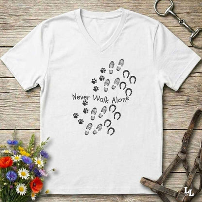 A white V-neck t-shirt with a black graphic print of paw prints, hoof prints, and the text 'Never Walk Alone' laid flat on a rustic wooden background with equestrian accessories and flowers.