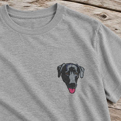 A close-up of a heather gray crewneck t-shirt featuring a stylized embroidered or printed graphic of a black and gray dog with its pink tongue out, laid flat on a rustic wooden surface.