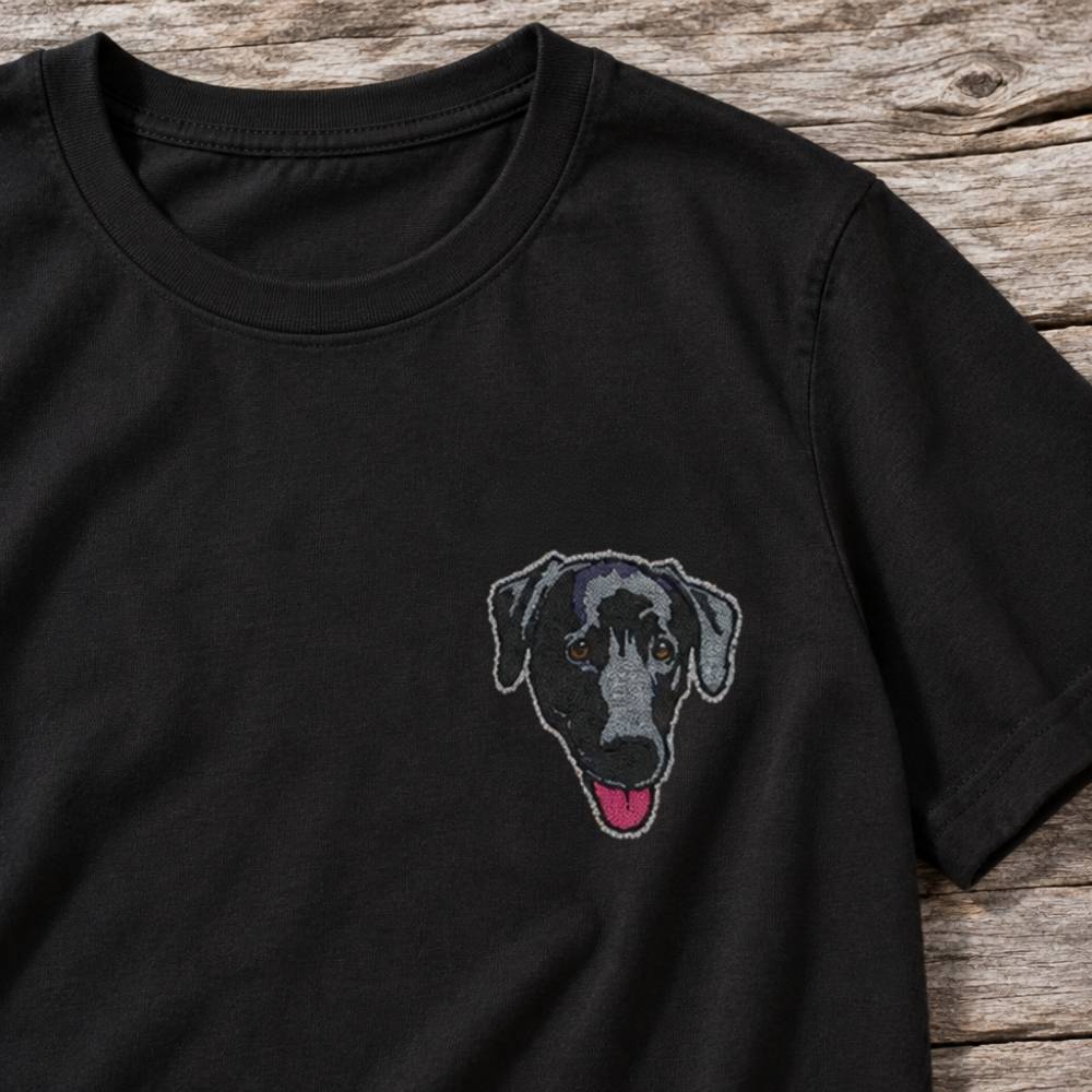 A black short-sleeve crewneck t-shirt featuring a custom embroidered dog face on the left chest, laid flat on a rustic wooden surface.