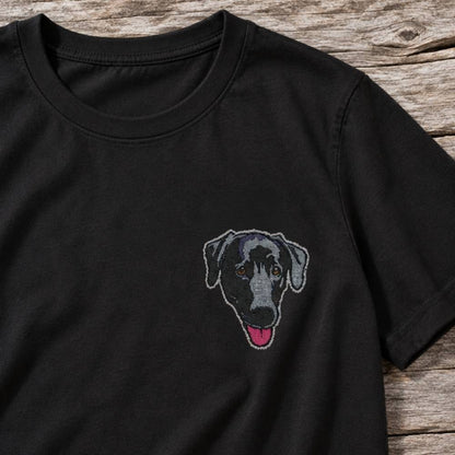 A black short-sleeve crewneck t-shirt featuring a custom embroidered dog face on the left chest, laid flat on a rustic wooden surface.