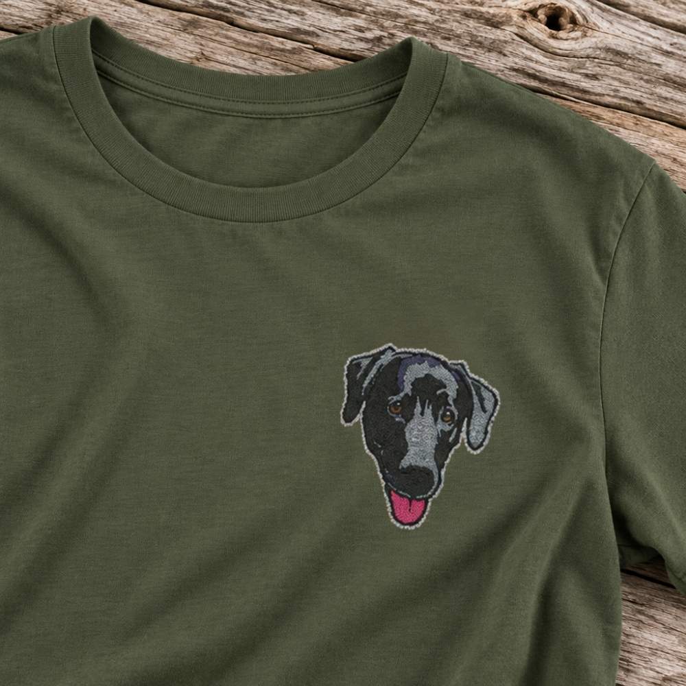 An olive green crewneck t-shirt featuring a detailed embroidered dog face on the left chest, laid flat on a rustic wooden surface.