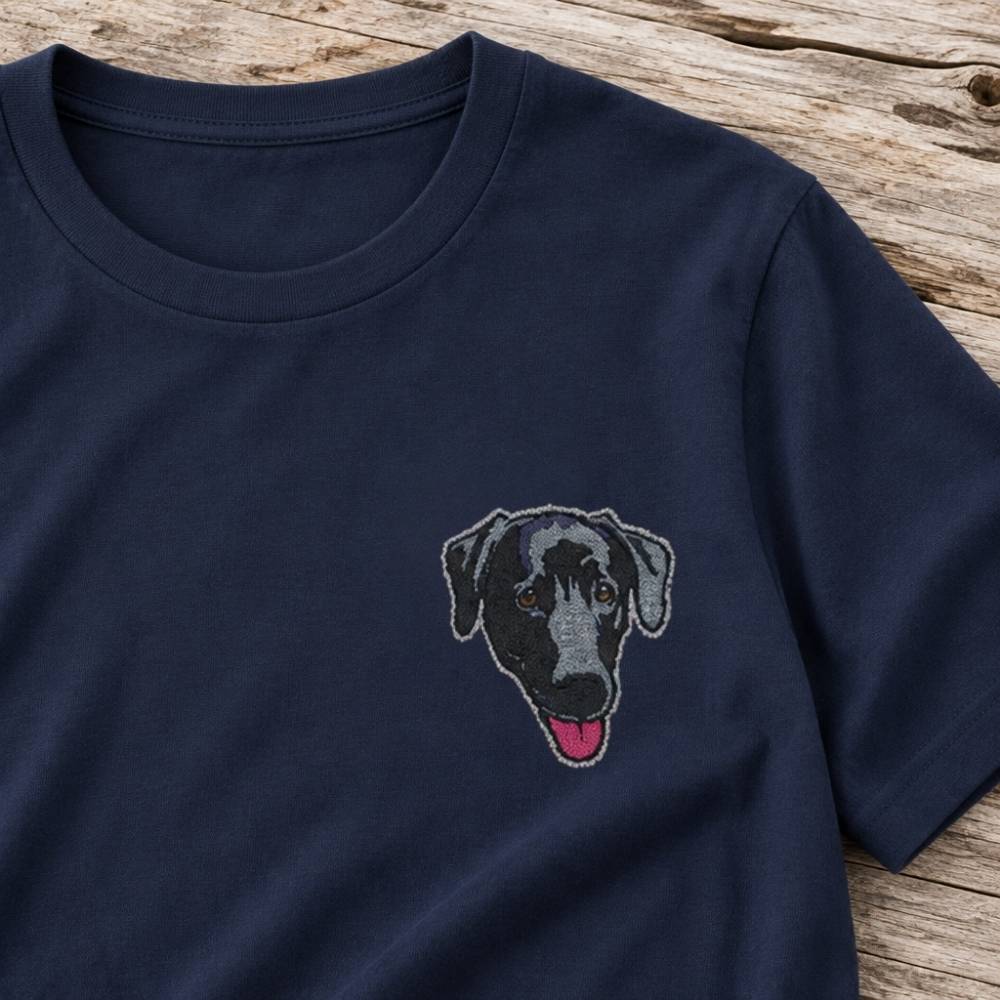 A close-up of a navy blue short-sleeve crewneck t-shirt featuring a custom embroidered dog face on the left chest, laid flat on a rustic wooden surface.