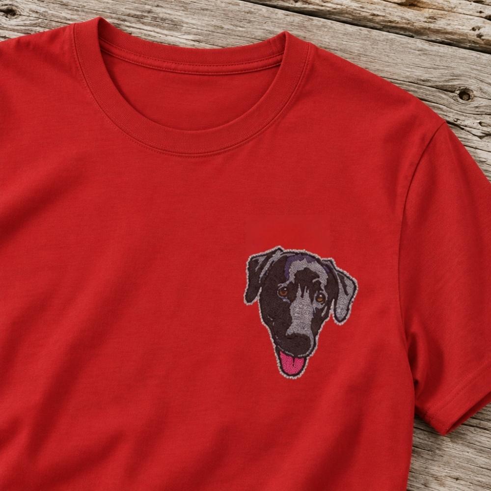 A red short-sleeve crewneck t-shirt featuring a detailed embroidered dog face on the left chest, laid flat on a rustic wooden surface.
