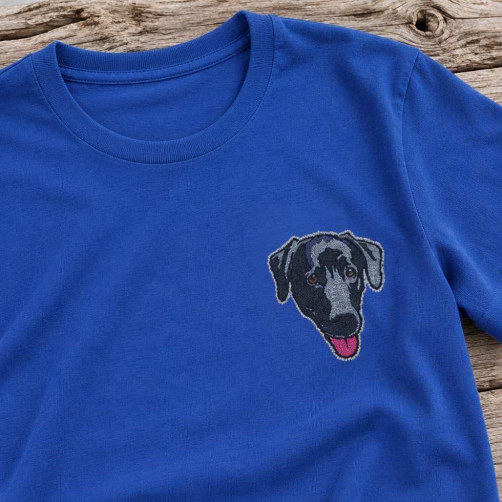A royal blue short-sleeve crewneck t-shirt featuring a detailed embroidered dog face on the left chest, laid flat on a rustic wooden surface.