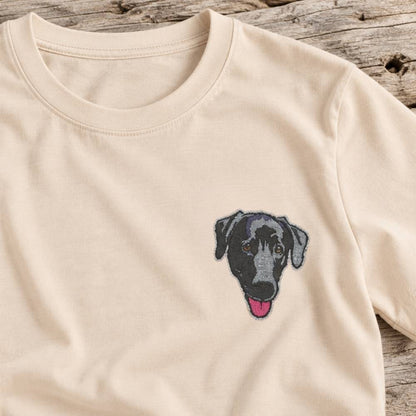 A close-up of a cream-colored crewneck t-shirt featuring a small embroidered black and gray dog face with a pink tongue on the left chest, laid flat on a rustic weathered wood background.