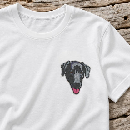 A white short-sleeve crewneck t-shirt featuring a detailed embroidered graphic of a black and gray dog's face with its pink tongue sticking out, laid flat on a rustic wooden plank background.