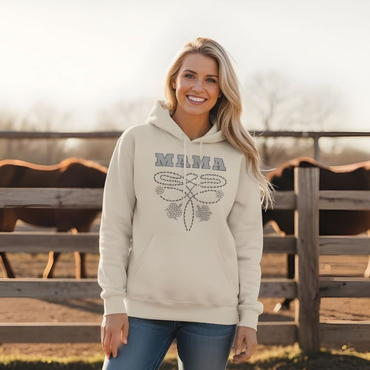 Woman wearing a beige hoodie with 'MAMA' and a bow design, standing in front of a wooden fence with horses.