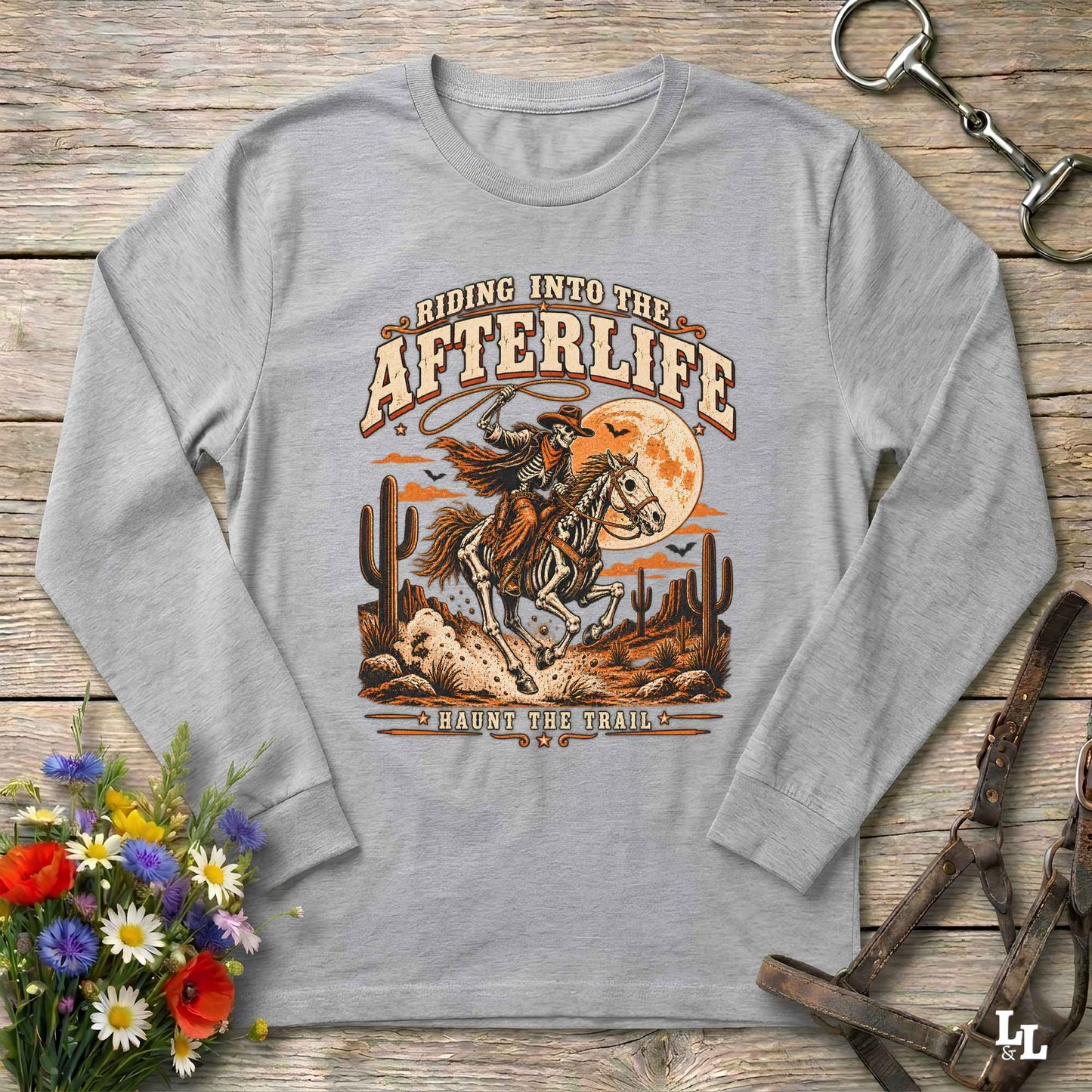 A light heather gray long-sleeve t-shirt with a vintage western graphic print, laid flat on a rustic wooden background with equestrian and floral props.