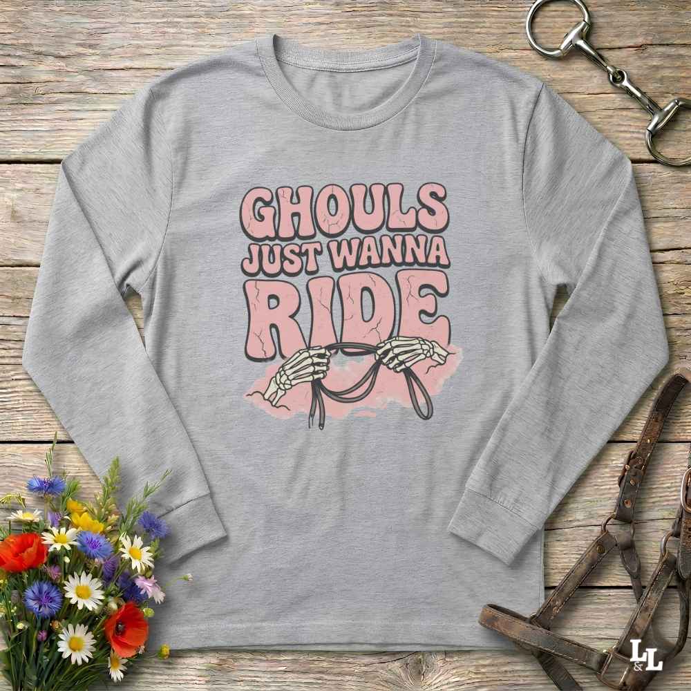 A light gray long-sleeve t-shirt with a graphic print reading 'GHOULS JUST WANNA RIDE' in pink and brown lettering, featuring skeleton hands holding reins. The shirt is displayed flat on a rustic wooden background with decorative props including a bouquet of flowers and horse tack.