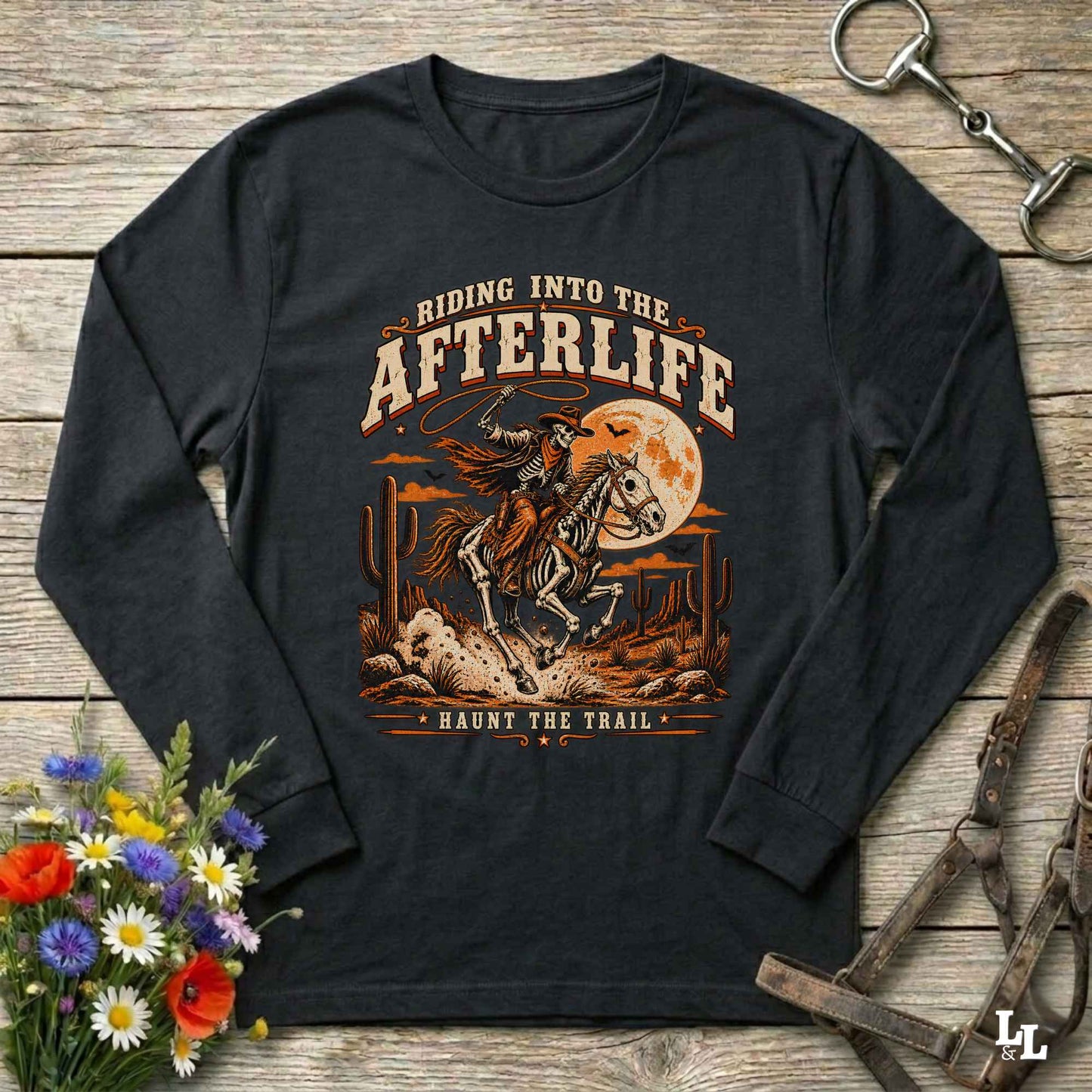 A black long-sleeve t-shirt with a western-themed graphic print, laid flat on a rustic wooden background with decorative props including a bouquet of flowers, a horse bit, and a leather bridle.