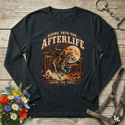 A black long-sleeve t-shirt with a western-themed graphic print, laid flat on a rustic wooden background with decorative props including a bouquet of flowers, a horse bit, and a leather bridle.