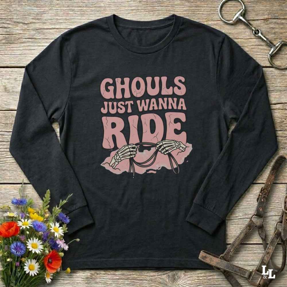 A black long-sleeve t-shirt with a pink graphic print that reads 'GHOULS JUST WANNA RIDE' above skeleton hands holding a horse bit, styled in a flat lay on a rustic wooden background with a horse bridle and flowers.