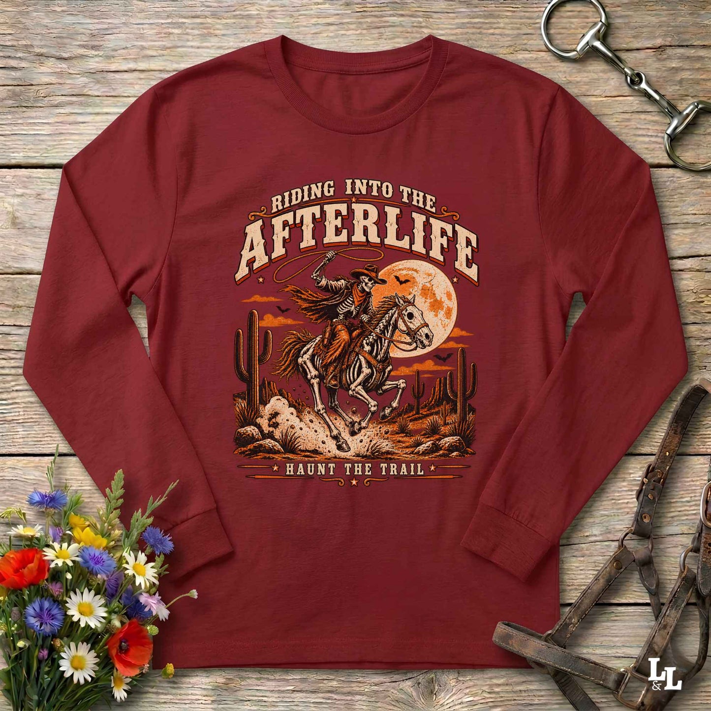 A long-sleeve crewneck t-shirt in maroon red, laid flat on a rustic wooden surface. The shirt features a detailed graphic print of a cowboy riding a horse across a desert landscape under a large orange full moon, with the text 'RIDING INTO THE AFTERLIFE' and 'HAUNT THE TRAIL'.