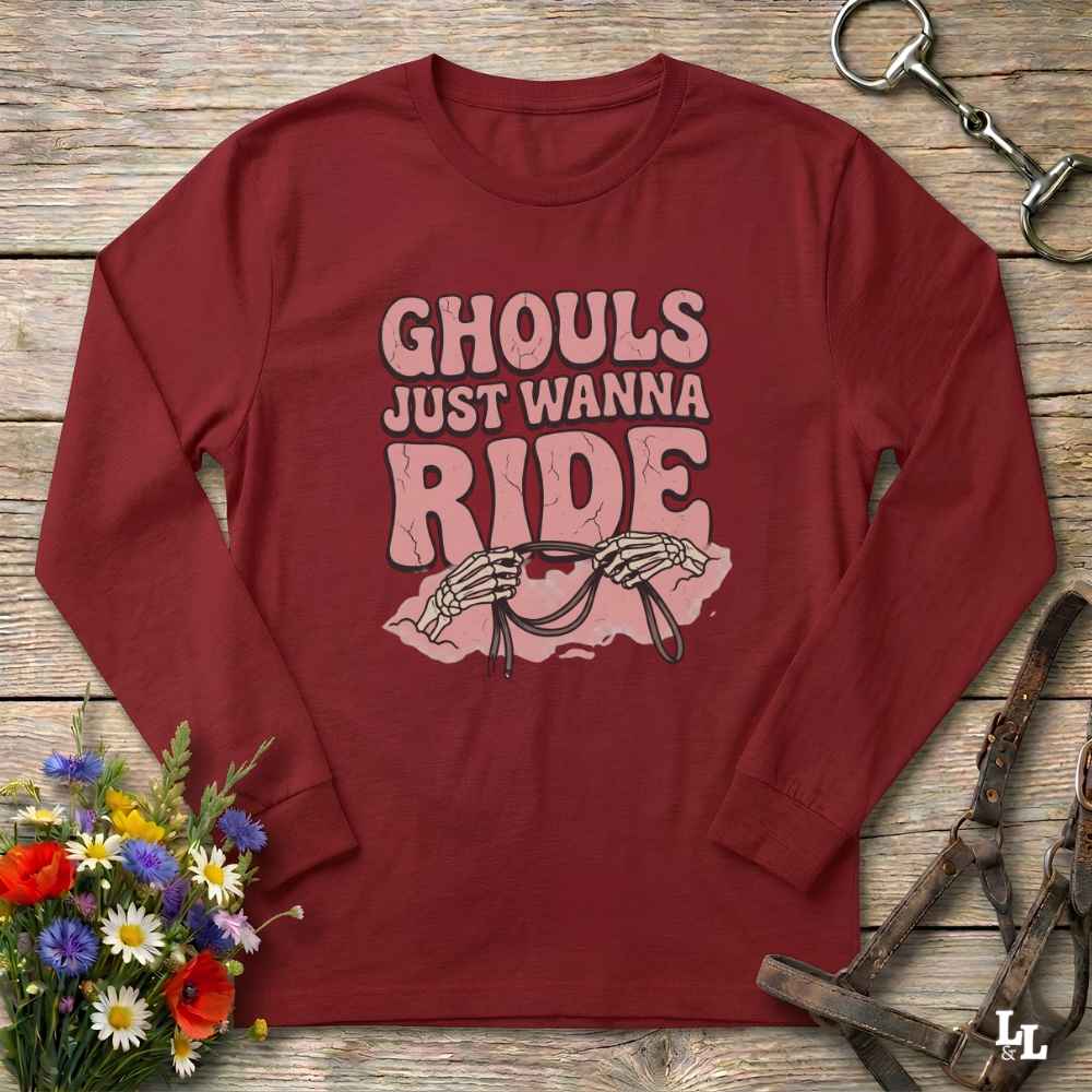 A maroon long-sleeve t-shirt with a pink graphic print that reads 'GHOULS JUST WANNA RIDE' above skeleton hands holding a horse bit, styled in a flat lay on a rustic wooden background with a horse bridle and flowers.