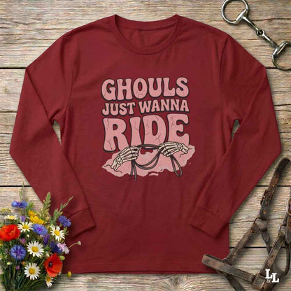 A maroon long-sleeve t-shirt with a pink graphic print that reads 'GHOULS JUST WANNA RIDE' above skeleton hands holding a horse bit, styled in a flat lay on a rustic wooden background with a horse bridle and flowers.