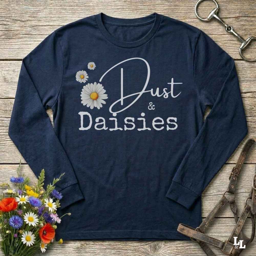 A navy blue long-sleeve t-shirt with a graphic print reading 'Dust & Daisies' in white script and block lettering, decorated with white and yellow daisy flowers. The shirt is displayed in a flat-lay arrangement on a rustic wooden background, surrounded by equestrian-themed props including a horse bit, leather bridle, and a small bouquet of wildflowers.