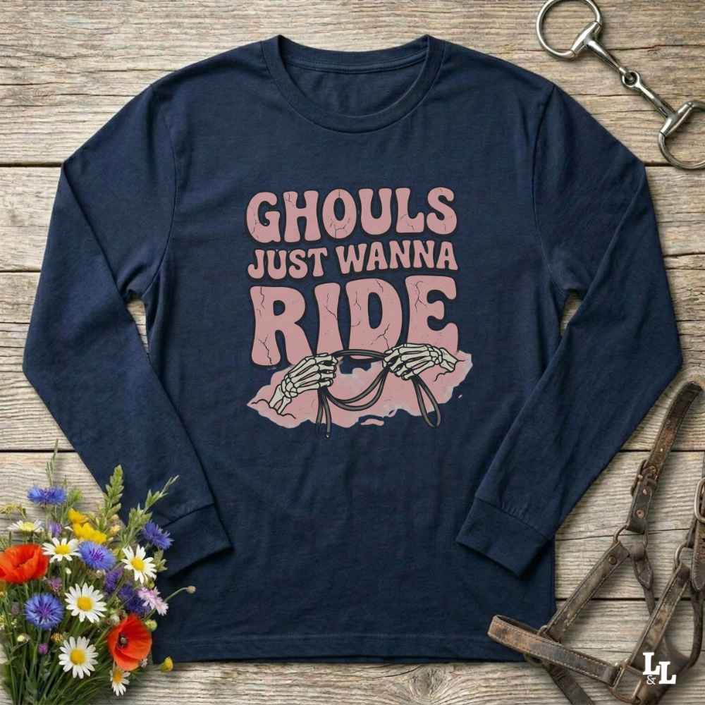 A navy blue long-sleeve t-shirt with a pink graphic print that reads 'GHOULS JUST WANNA RIDE' above skeleton hands holding a rearing horse. The shirt is displayed in a flat-lay arrangement on a rustic wooden background, surrounded by decorative props including a bouquet of flowers, a horse bit, and a leather bridle.