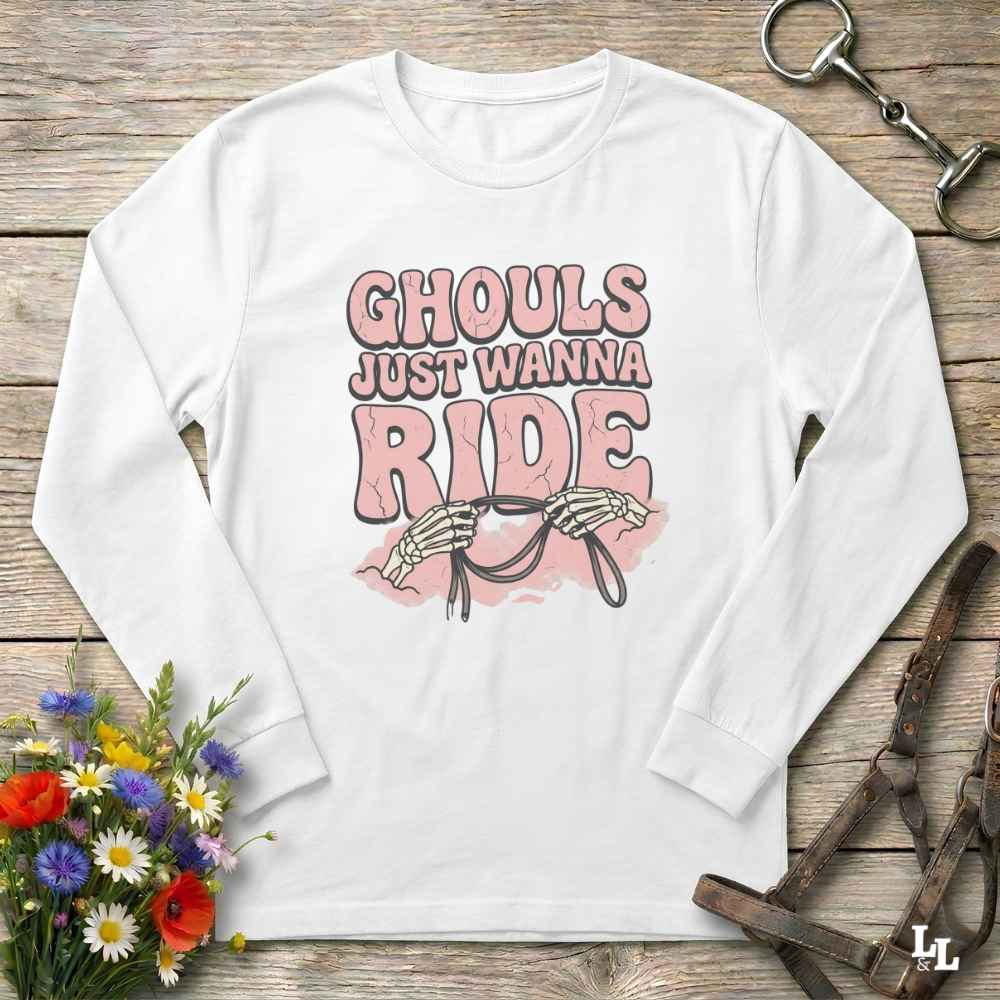 A white long-sleeve t-shirt with a graphic print reading 'GHOULS JUST WANNA RIDE' in pink and black, featuring skeleton hands holding a bridle. The shirt is laid flat on a rustic wooden background with equestrian-themed props like a leather bridle and a bouquet of wildflowers.