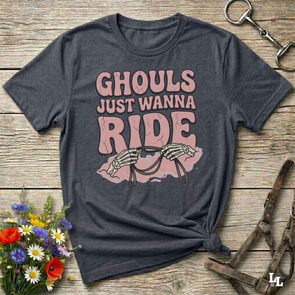 A grey short-sleeve graphic T-shirt laid flat on a rustic wooden surface, with the text 'GHOULS JUST WANNA RIDE' and a skeleton riding a horse design. Decorative flowers and horse tack are placed around it.