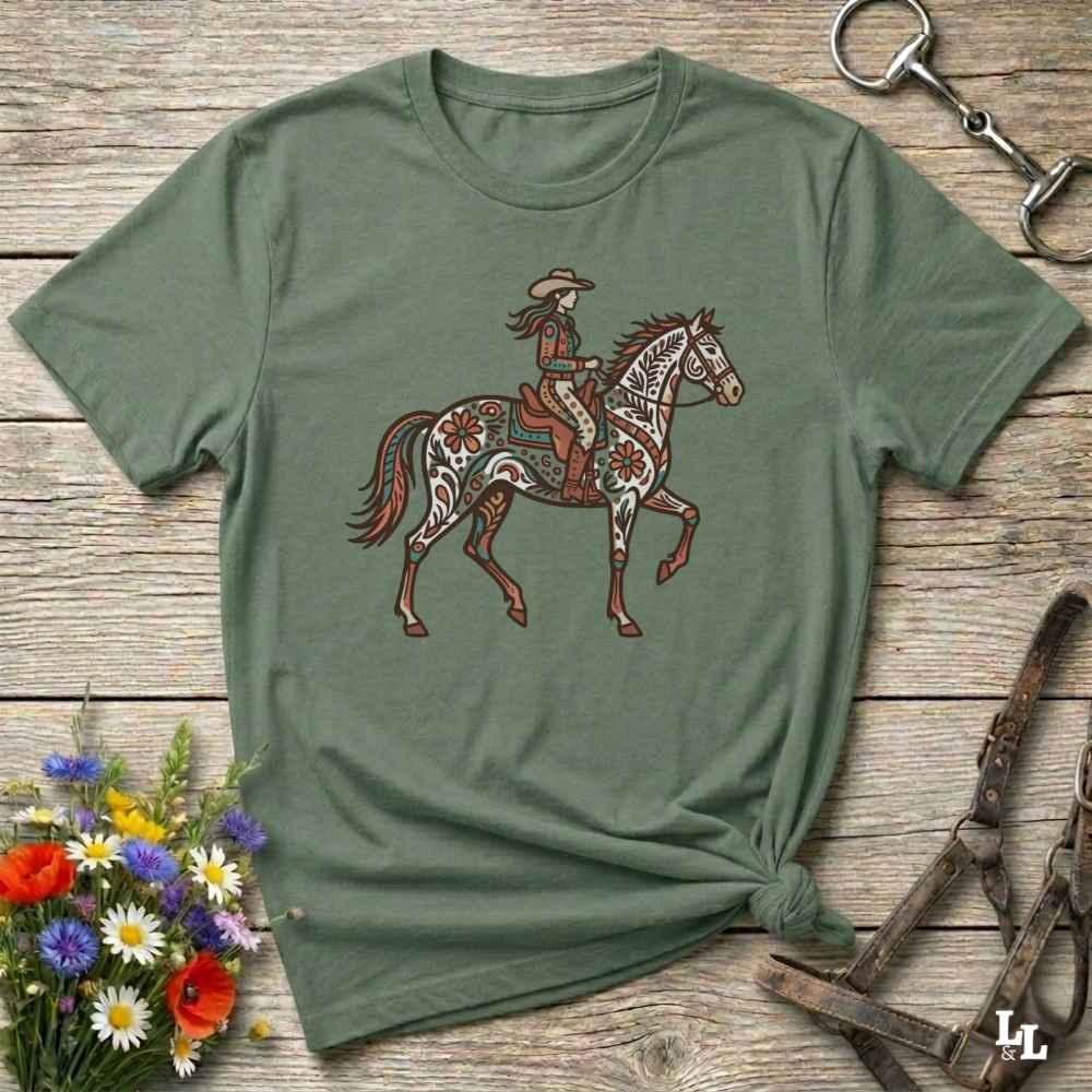 Green t-shirt with a graphic of a woman riding a horse on a wooden surface with floral and equestrian-themed accessories.
