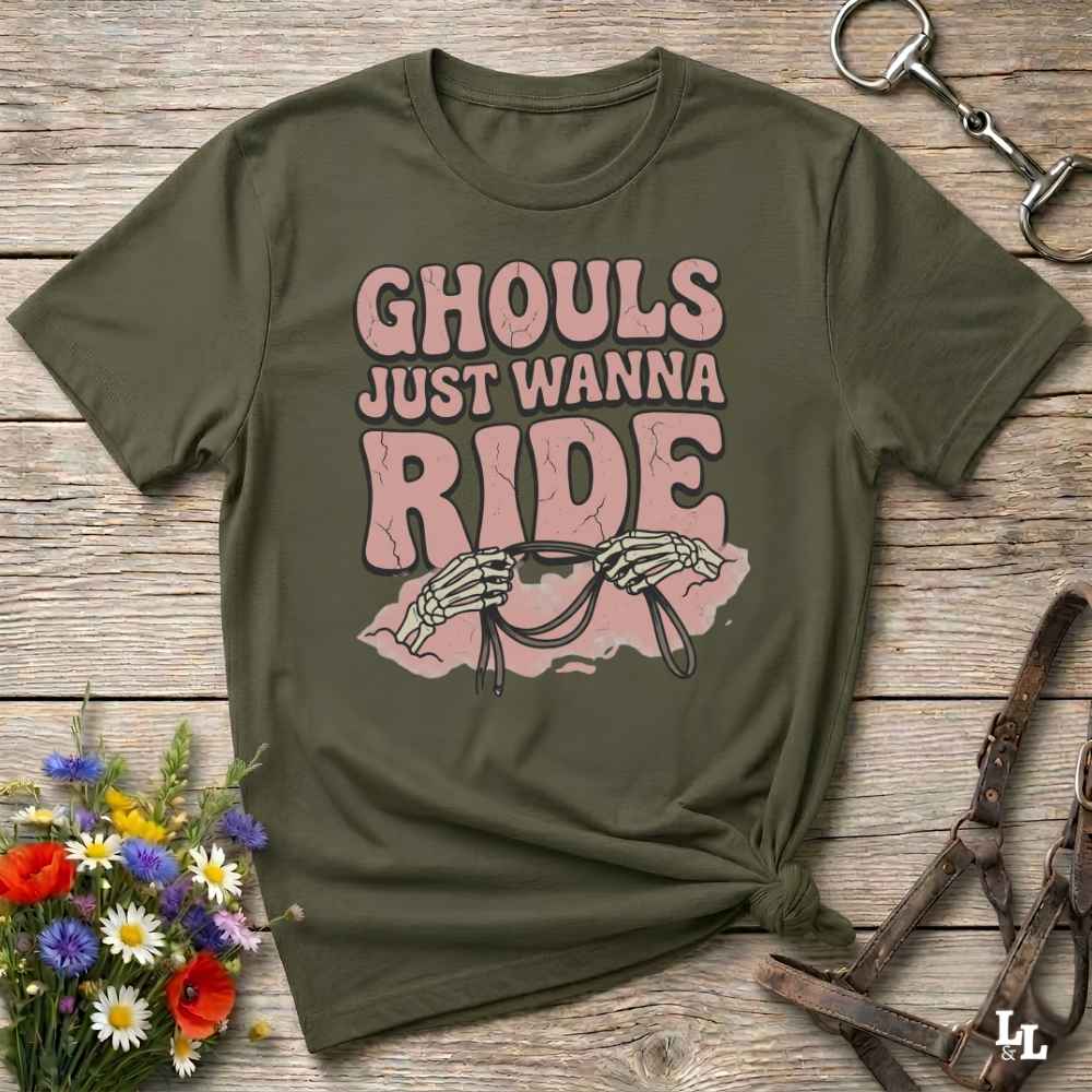 An olive green graphic t-shirt laid flat on a rustic wooden surface, styled with a small bouquet of flowers, a horse bit, and a leather bridle. The t-shirt features a pink graphic that reads 'GHOULS JUST WANNA RIDE' with skeleton hands holding reins.