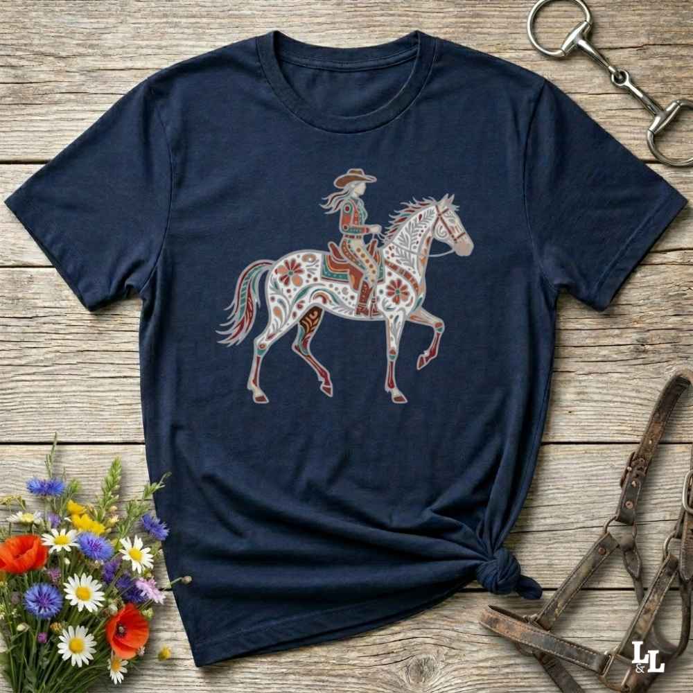 Navy blue t-shirt with a colorful horse and rider graphic on a wooden surface with flowers and equestrian tools.