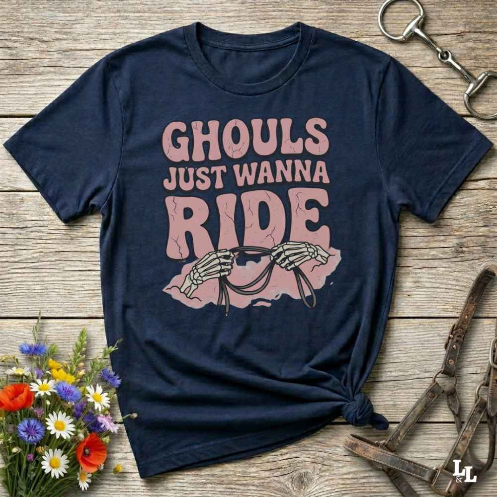 A navy blue graphic t-shirt laid flat on a rustic wooden surface, styled with equestrian props like a metal horse bit, a leather bridle, and a small bouquet of wildflowers. The shirt features a pink graphic that reads 'GHOULS JUST WANNA RIDE' with skeleton hands holding reins.