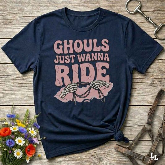 A navy blue graphic t-shirt laid flat on a rustic wooden surface, styled with equestrian props like a metal horse bit, a leather bridle, and a small bouquet of wildflowers. The shirt features a pink graphic that reads 'GHOULS JUST WANNA RIDE' with skeleton hands holding reins.