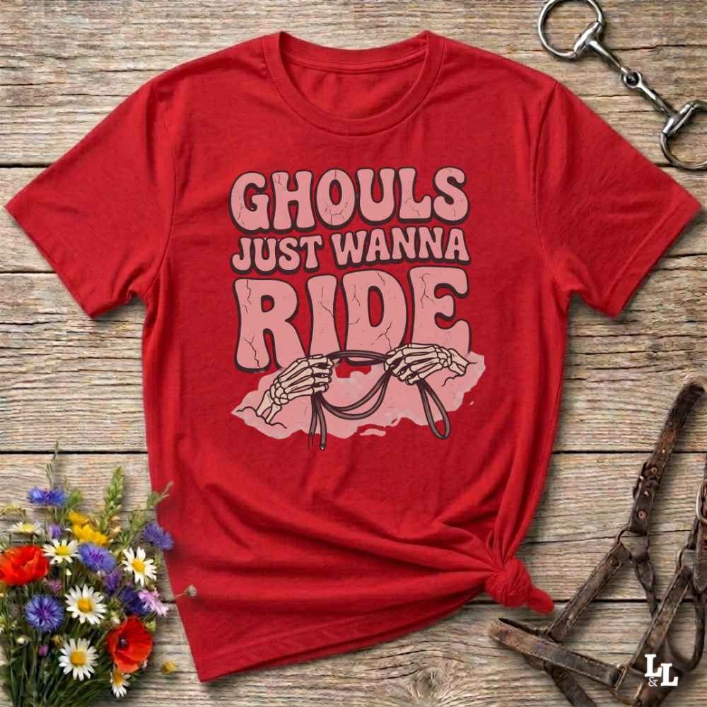 A red short-sleeve graphic T-shirt laid flat on a rustic wooden surface, with the text 'GHOULS JUST WANNA RIDE' and a skeleton riding a horse design. Decorative flowers and horse tack are placed around it.