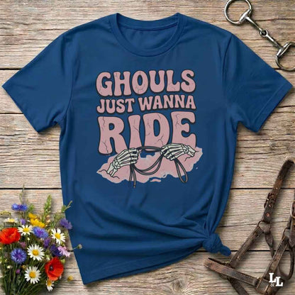 A blue short-sleeve graphic T-shirt laid flat on a rustic wooden surface, with the text 'GHOULS JUST WANNA RIDE' and a skeleton riding a horse design. Decorative flowers and horse tack are placed around it.