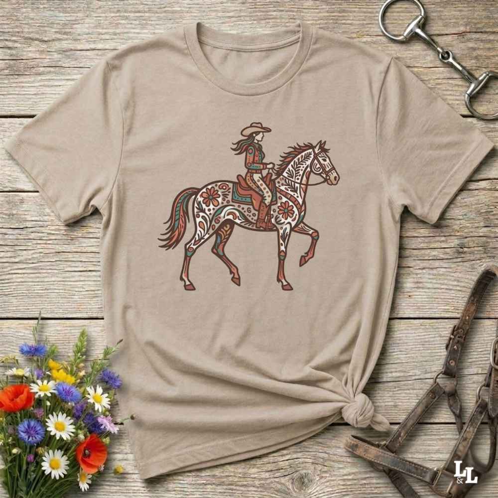 Beige t-shirt with a colorful graphic of a woman riding a horse on a wooden surface with floral and equestrian-themed accessories.
