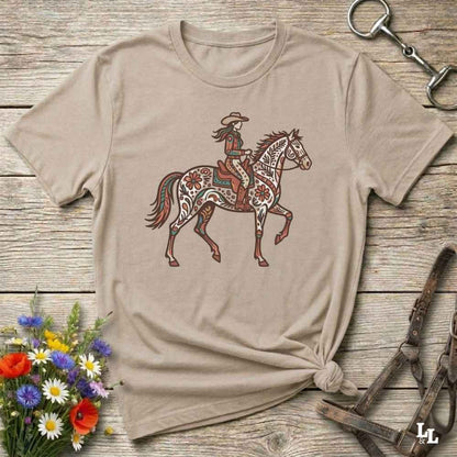 Beige t-shirt with a colorful graphic of a woman riding a horse on a wooden surface with floral and equestrian-themed accessories.