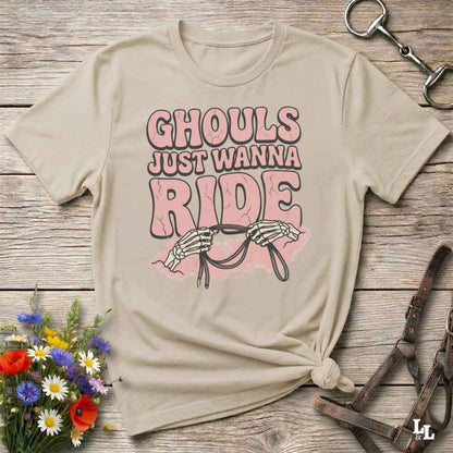 A beige graphic t-shirt laid flat on a rustic wooden surface, styled with a small bouquet of wildflowers, a horse bit, and a leather bridle. The t-shirt features a graphic print that reads 'GHOULS JUST WANNA RIDE' in pink and dark brown lettering, accompanied by a skeleton holding reins.